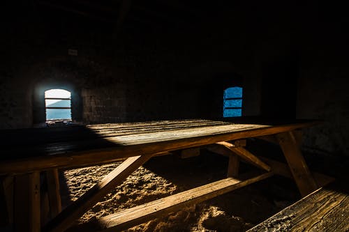 Empty picnic table silhouetted with two windows in the background on a dirt floor.