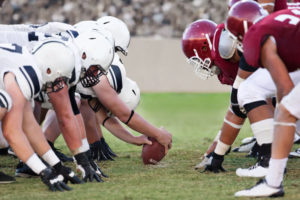 Two football teams line up on the scrimmage line. The team in white is on offense. The team in red is on defense.