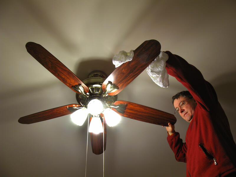 Man wearing red sweatshirt wiping down bedroom ceiling fan with a rag.