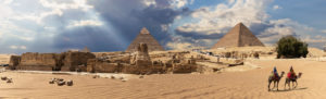 Two people on camels cross a pyramid burial complex.